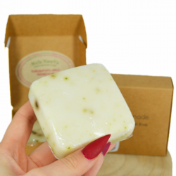 Hand holding a pale soap bar with herb pieces, cardboard box and packaging in background